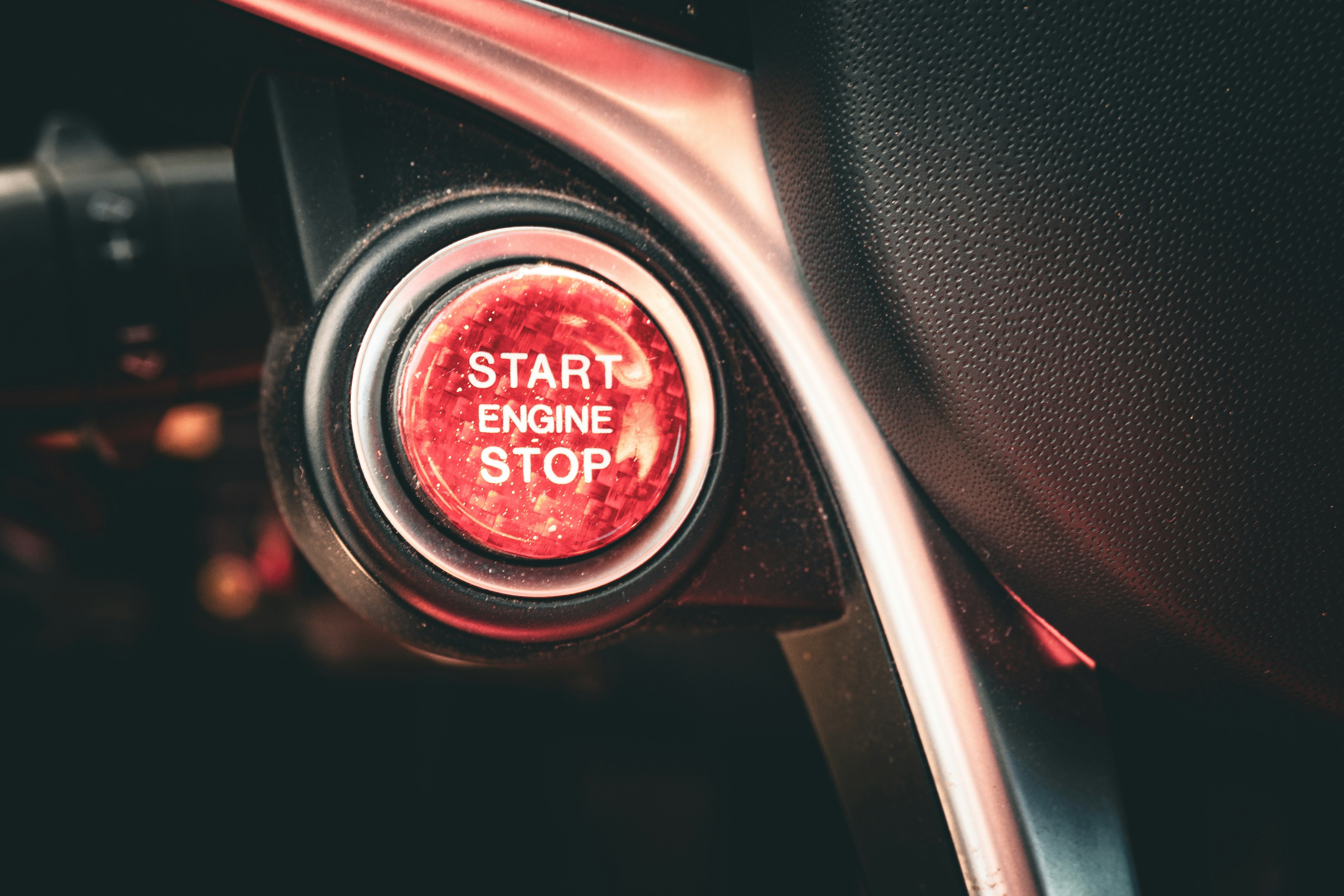 A red carbon-textured engine start/stop button glowing in a dark car interior. Macro detail of the \